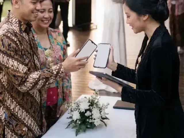 Tamu berbatik menunjukkan QR di HP kepada penerima tamu yang memindainya sambil memegang tablet.